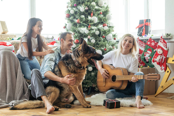 χαμογελαστή οικογένεια και κόρη με το σκυλί κάθεται κοντά στο χριστουγεννιάτικο δέντρο με δώρα. - Φωτογραφία, εικόνα