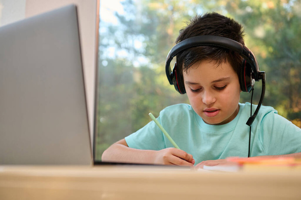 Close-up of smart concentrated teenage schoolboy wearing audio headset, sitting at a desk with laptop and making notes during the online lesson with remote web teacher. Homeschooling. Online education - 写真・画像