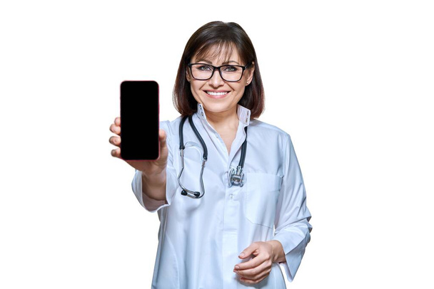 Woman doctor in white uniform with stethoscope showing smartphone screen to camera, on white isolated background. Medicine technology applications health care, mockup banner telemedicine - Фото, зображення