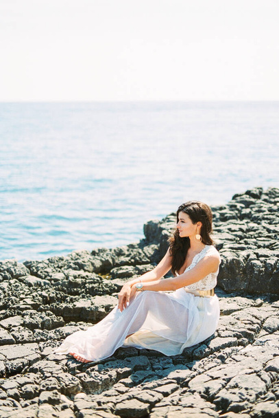 Наречена босоніж у білій сукні сидить на скелі біля моря і дивиться на воду. Високоякісна фотографія
 - Фото, зображення