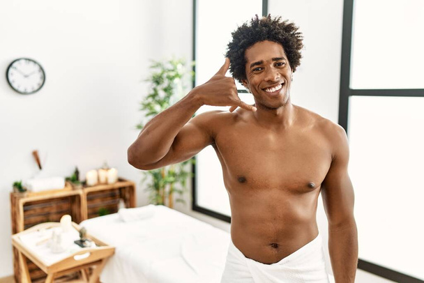 Jeune homme afro-américain torse nu portant une serviette debout au centre de beauté souriant faire geste de téléphone avec la main et les doigts comme parler au téléphone. communiquer les concepts.  - Photo, image