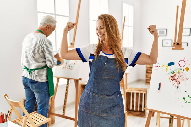 Hispanic woman wearing apron at art studio showing arms muscles smiling proud. fitness concept.  - Foto, Imagen