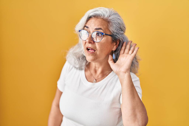 Μεσήλικη γυναίκα με γκρίζα μαλλιά στέκεται πάνω από το κίτρινο φόντο χαμογελώντας με το χέρι πάνω από το αυτί ακούγοντας μια ακρόαση για φήμες ή κουτσομπολιά. έννοια της κώφωσης.  - Φωτογραφία, εικόνα