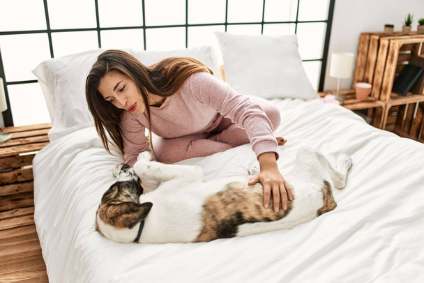 Молодая женщина улыбается уверенно играя с собакой сидя на кровати в спальне - Фото, изображение