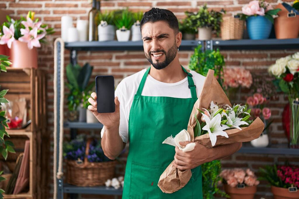 Joven hispano que trabaja en floristería mostrando la pantalla del teléfono inteligente sin pistas y expresión confusa. concepto de duda.  - Foto, imagen