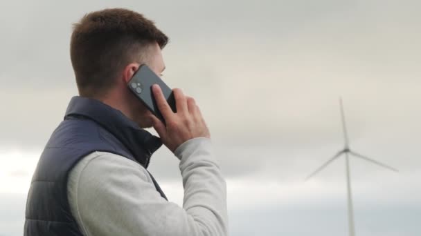 Erkek mühendis rüzgar türbininin rüzgar değirmenine bakan akıllı telefondan çalıştığını rapor ediyor. Rüzgar türbini gri gökyüzüne karşı yeşil enerji üretir - Video, Çekim