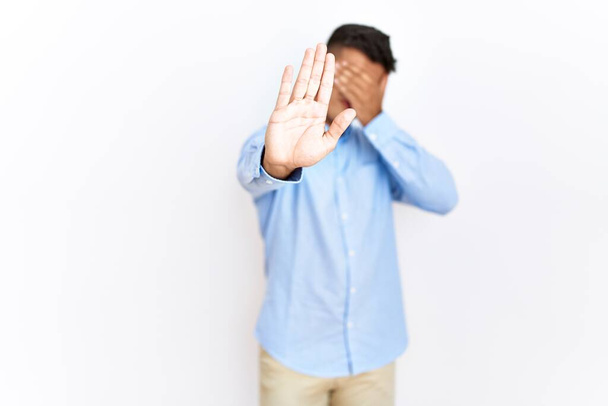 Jeune homme hispanique portant une chemise d'affaires debout sur un fond isolé couvrant les yeux des mains et faisant un geste d'arrêt avec expression triste et peur. concept embarrassé et négatif.  - Photo, image