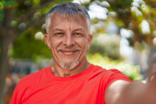 Grauhaariger Mann mittleren Alters lächelt selbstbewusst und macht Selfie mit Kamera im Park - Foto, Bild