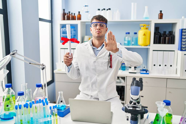 Молодой латиноамериканец, работающий в лаборатории ученого, держит диплом с открытой рукой и делает знак стоп с серьезным и уверенным выражением, защитный жест  - Фото, изображение