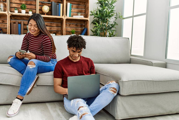 Junges lateinisches Paar lächelt glücklich mit Laptop und Smartphone zu Hause. - Foto, Bild