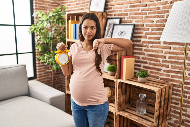 Young pregnant woman holding alarm clock for due date with angry face, negative sign showing dislike with thumbs down, rejection concept  - Foto, Imagen