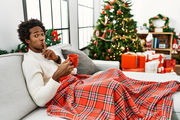Junger afrikanisch-amerikanischer Mann sitzt auf dem Sofa und trinkt Kaffee am Weihnachtsbaum und bittet um Ruhe mit dem Finger auf den Lippen. Schweigen und geheimes Konzept.  - Foto, Bild