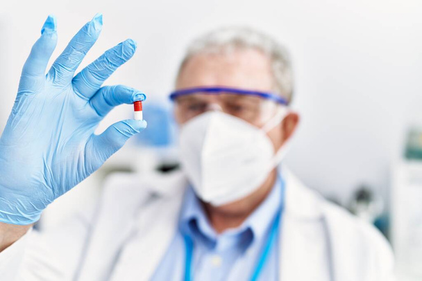 Middle age grey-haired man wearing scientist uniform hoding pill at laboratory - Photo, Image