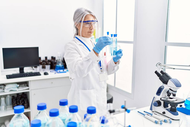 Mulher de cabelos grisalhos de meia idade usando uniforme cientista usando laptop segurando tubo de teste em laboratório - Foto, Imagem