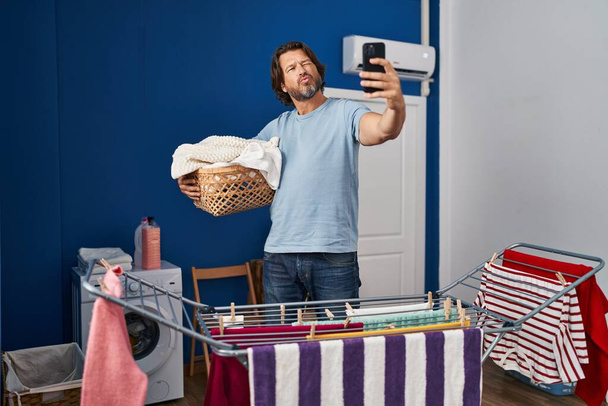 Handsome middle age man holding laundry basket doing selfie picture looking at the camera blowing a kiss being lovely and sexy. love expression.  - Photo, image