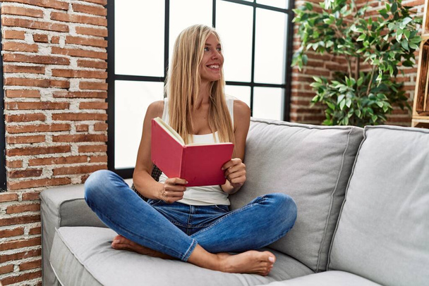 Young blonde woman reading book sitting on sofa at home - Foto, immagini