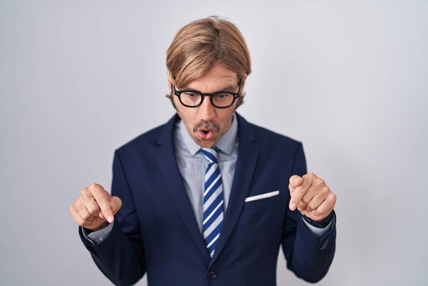 Caucasian man with mustache wearing business clothes pointing down with fingers showing advertisement, surprised face and open mouth  - Foto, Imagem
