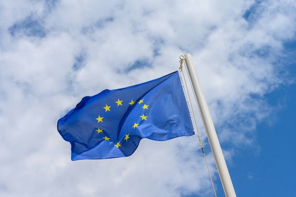Σημαία της Ευρωπαϊκής Ένωσης κυματίζει στον άνεμο. Είναι μια όμορφη ηλιόλουστη μέρα του καλοκαιριού, με μπλε ουρανό και λευκά σύννεφα στο παρασκήνιο. - Φωτογραφία, εικόνα