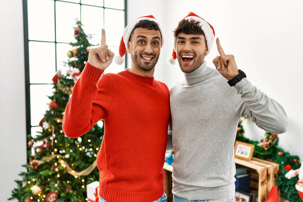Young gay couple standing by christmas tree wearing hat pointing finger up with successful idea. exited and happy. number one.  - Photo, Image