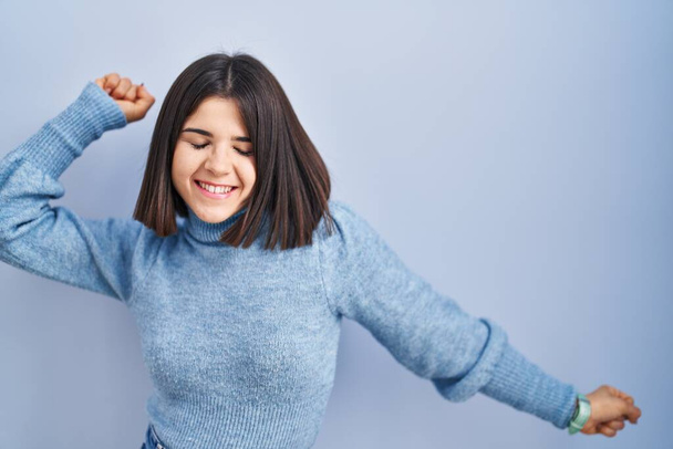 Young hispanic woman standing over blue background dancing happy and cheerful, smiling moving casual and confident listening to music  - Foto, Imagen
