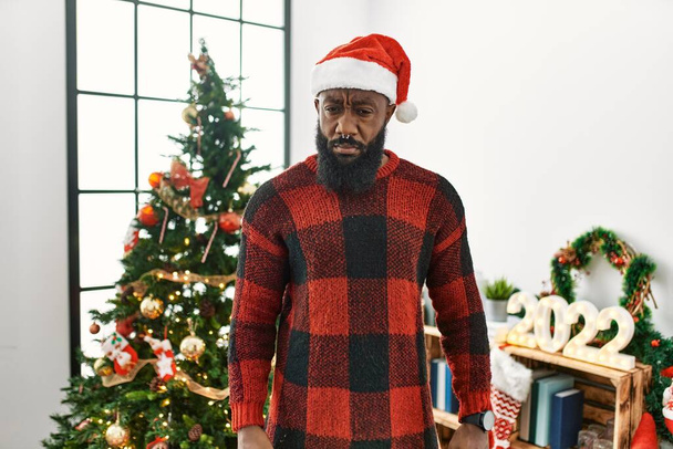 Αφροαμερικάνος που φοράει καπέλο Άη Βασίλη στέκεται δίπλα στο χριστουγεννιάτικο δέντρο σκεπτικός και νευρικός, συνοφρυωμένος λόγω προβλήματος. αρνητικό πρόσωπο.  - Φωτογραφία, εικόνα