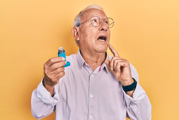 Senior man with grey hair holding medical asthma inhaler serious face thinking about question with hand on chin, thoughtful about confusing idea  - 写真・画像