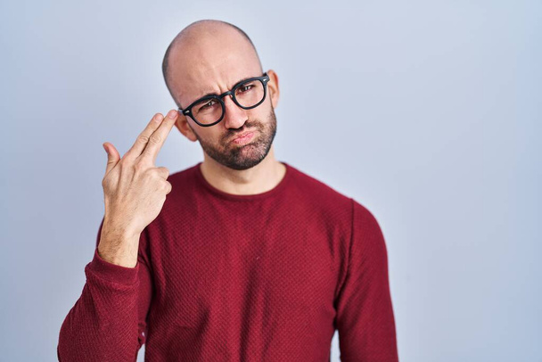 Young bald man with beard standing over white background wearing glasses shooting and killing oneself pointing hand and fingers to head like gun, suicide gesture.  - Foto, immagini