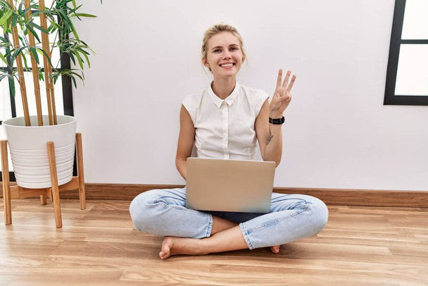 Jovem loira usando computador portátil sentado no chão na sala de estar mostrando e apontando para cima com os dedos número três, sorrindo confiante e feliz.  - Foto, Imagem