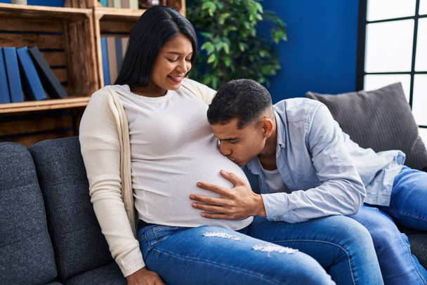 Young latin couple expecting baby kissing belly sitting on sofa at home - Fotó, kép
