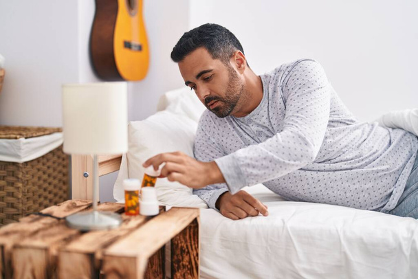 Joven hispano acostado en la cama sosteniendo pastillas en el dormitorio - Foto, Imagen