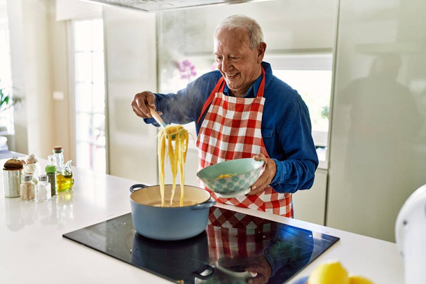 Hombre mayor sonriendo confiado cocinando espaguetis en la cocina - Foto, Imagen