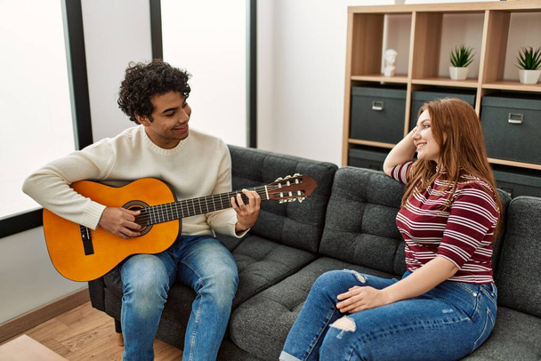 Młoda para uśmiechnięta szczęśliwa grając na gitarze klasycznej siedząc na kanapie w domu. - Zdjęcie, obraz