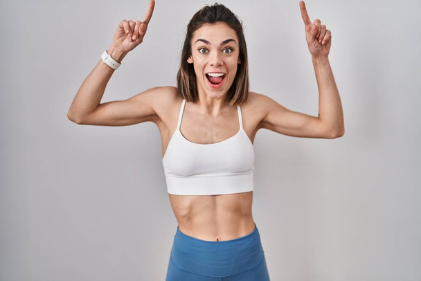 Femme hispanique portant des vêtements de sport sur fond isolé souriant étonné et surpris et pointant vers le haut avec les doigts et les bras levés.  - Photo, image