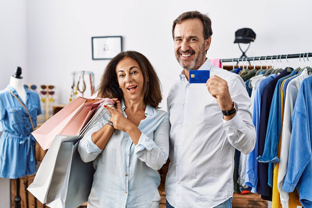 Hispanic middle age couple holding shopping bags and credit card with a big smile on face, pointing with hand finger to the side looking at the camera.  - Foto, imagen