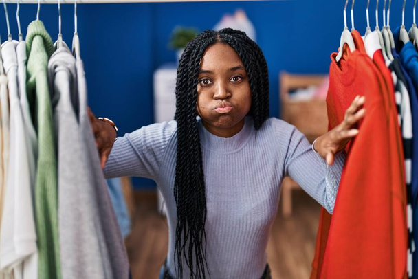 Afro-Amerikaanse vrouw op zoek naar kleren op kledingrek puffend wangen met grappig gezicht. mond opgeblazen met lucht, vangen van lucht.  - Foto, afbeelding