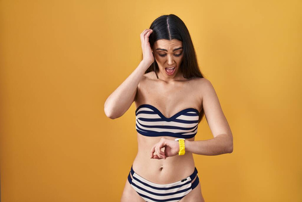 jonge brunette vrouw dragen bikini over gele achtergrond kijken naar het horloge tijd bezorgd, bang voor het krijgen van laat  - Foto, afbeelding