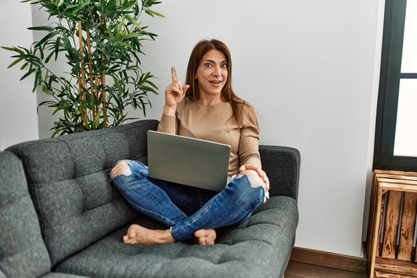 Végzős közel-keleti nő laptopot használ, otthon ül a kanapén, ujjal mutogatva a sikeres ötletre. kilépett és boldog volt. Egyes számú..  - Fotó, kép
