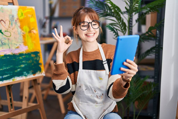 Νεαρή όμορφη καλλιτέχνης γυναίκα κάνει βιντεοκλήση με tablet κάνει ok υπογράψει με τα δάχτυλα, χαμογελώντας φιλικό gesturing εξαιρετικό σύμβολο  - Φωτογραφία, εικόνα