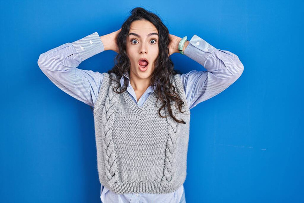 Young brunette woman standing over blue background crazy and scared with hands on head, afraid and surprised of shock with open mouth  - Photo, Image