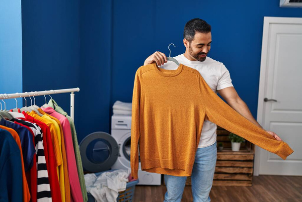 Young hispanic man holding sweater at laundry room - Foto, afbeelding