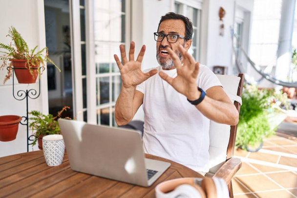 Middle age man using computer laptop at home afraid and terrified with fear expression stop gesture with hands, shouting in shock. panic concept.  - 写真・画像