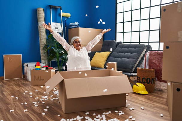 Mujer de mediana edad sonriendo confiado lanzando paquete de espuma en el nuevo hogar - Foto, Imagen
