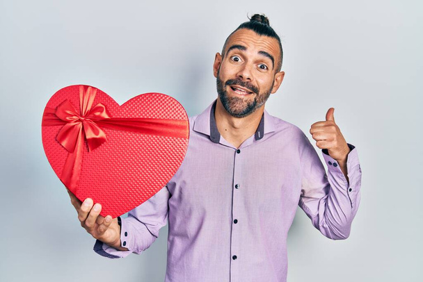 Junger hispanischer Mann mit einem Valentinstagsgeschenk, das den Daumen zur Seite zeigt und glücklich mit offenem Mund lächelt  - Foto, Bild
