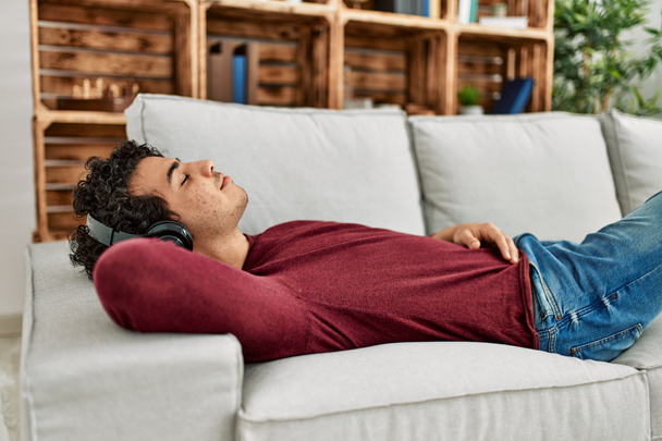 Joven hombre hispano relajado escuchando música tumbado en el sofá en casa. - Foto, imagen