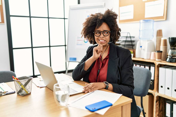 Afro-Amerikaanse vrouw met afrokapsel werkend op kantoor met operatorheadset denkend aan een vraag, bezorgd en nerveus met de hand op de kin  - Foto, afbeelding