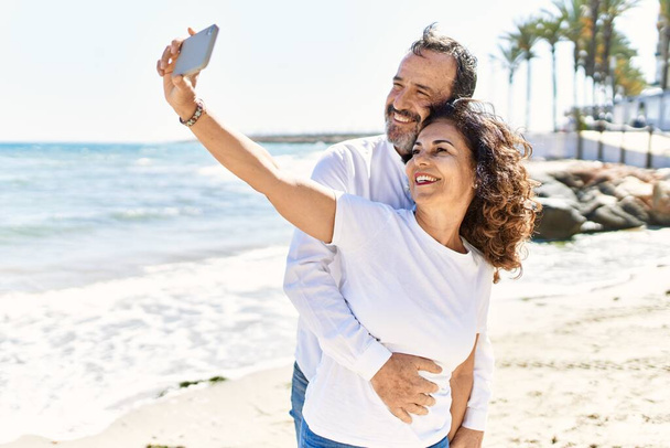 Middelbare leeftijd hispanic paar glimlachen gelukkig en knuffelen maken selfie door de smartphone op het strand. - Foto, afbeelding