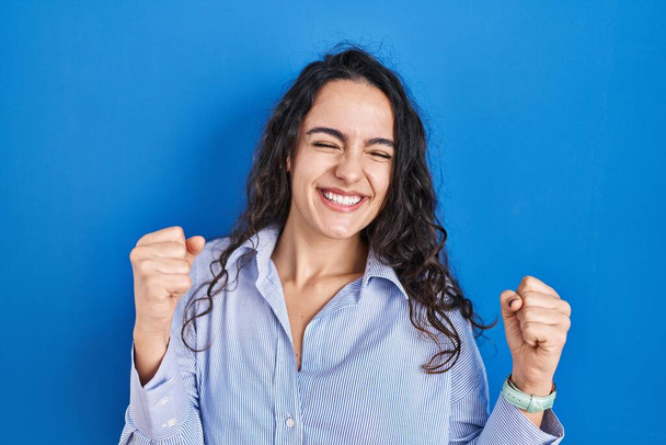 青の背景に立って若いブルネットの女性は非常に幸せと腕を上げて勝者のジェスチャーを行う興奮し、笑顔と成功のために叫んでいます。お祝いのコンセプト.  - 写真・画像