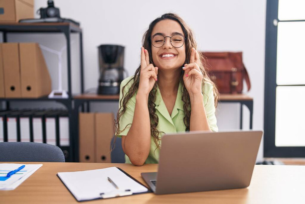 Junge hispanische Frau, die im Büro arbeitet, mit Brille, erhobenen Zeigefinger, lächelnd mit Hoffnung und geschlossenen Augen. Glück und abergläubisches Konzept.  - Foto, Bild