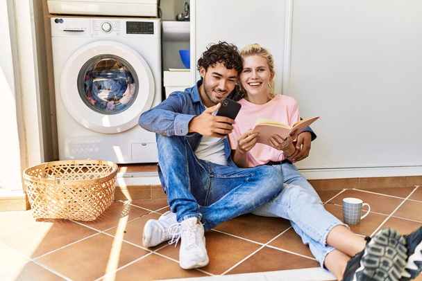 Young couple smiling happy reading book and using smartphone while doing laundry at home. - Photo, Image