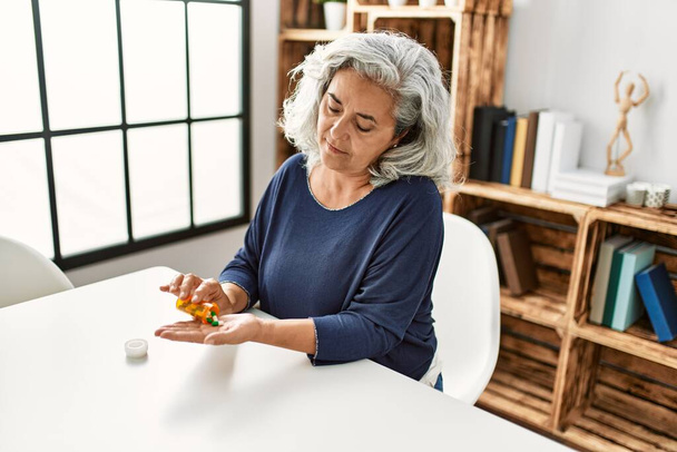 Middle age grey-haired woman taking pills sitting on the table at home. - Foto, Bild
